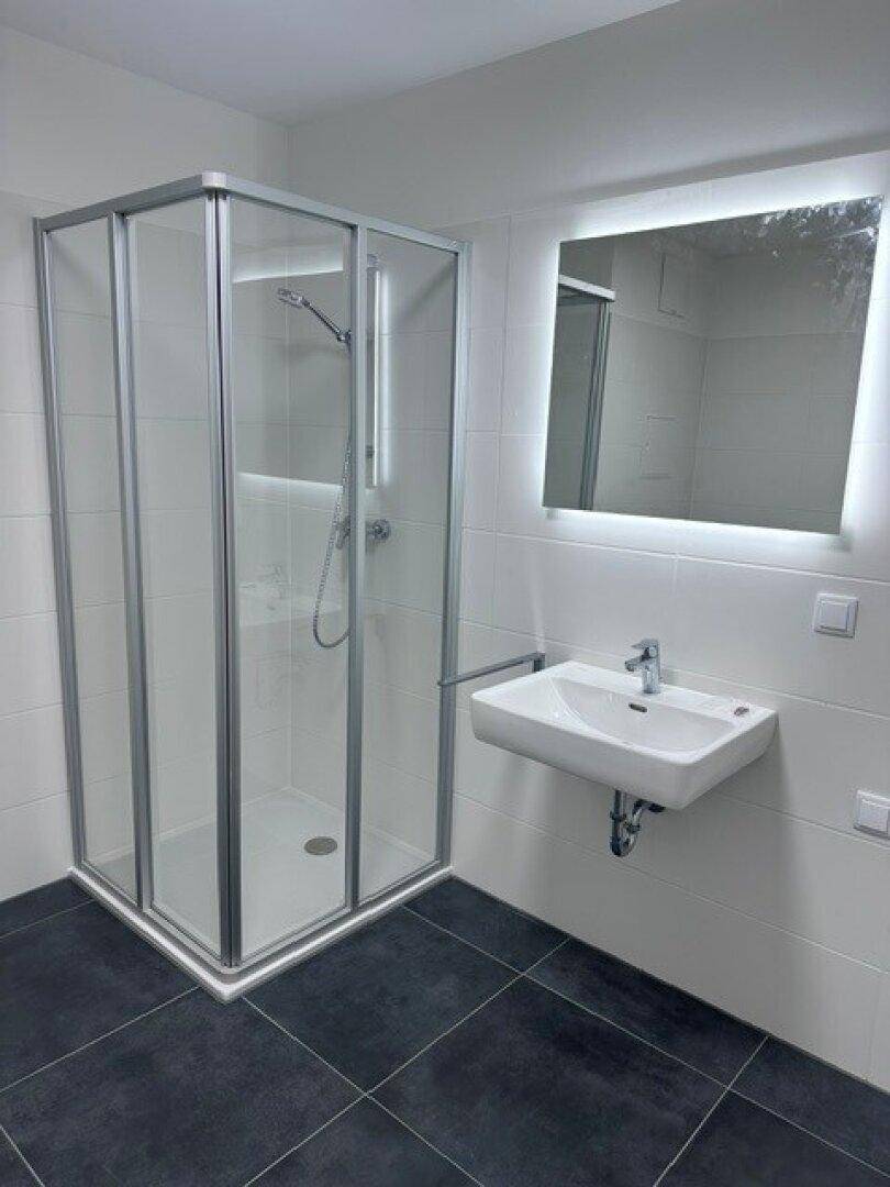 Helles Badezimmer mit Duschkabine, wandhängendem Waschbecken und beleuchtetem Spiegel.