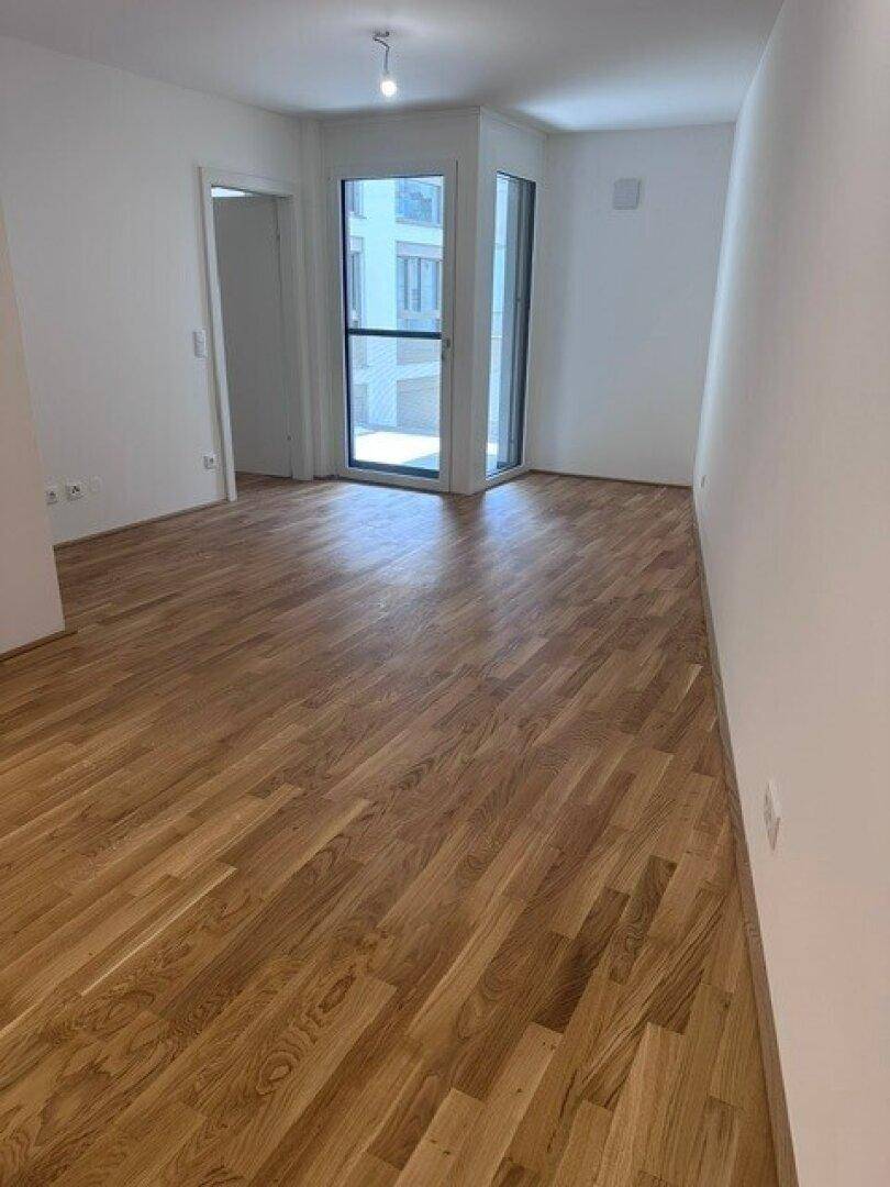 Heller Raum mit Parkettboden und großen Fenstern, die Zugang zu einem Außenbereich bieten.