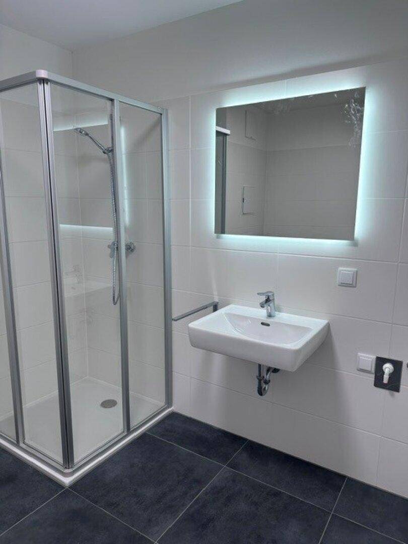 Modernes Badezimmer mit Duschkabine, beleuchtetem Spiegel und dunklen Bodenfliesen.