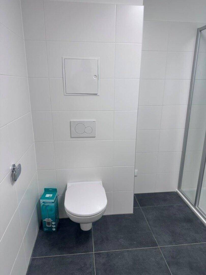 Separate Toilette mit Wand-WC und weißen Fliesen, angrenzend an eine Duschkabine.
