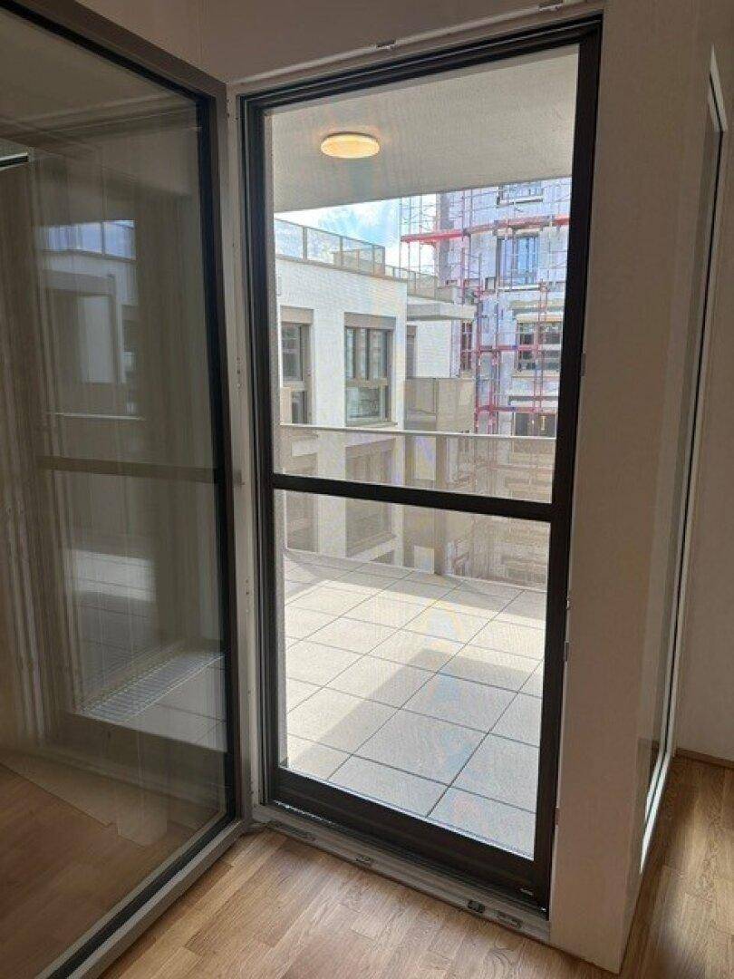 Innenansicht eines Zimmers mit großem Fenster und Zugang zu einem gefliesten Balkon.