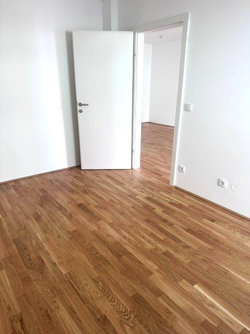 Leerer Raum mit Holzboden und offener Tür, der auf weitere Zimmer blicken lässt.