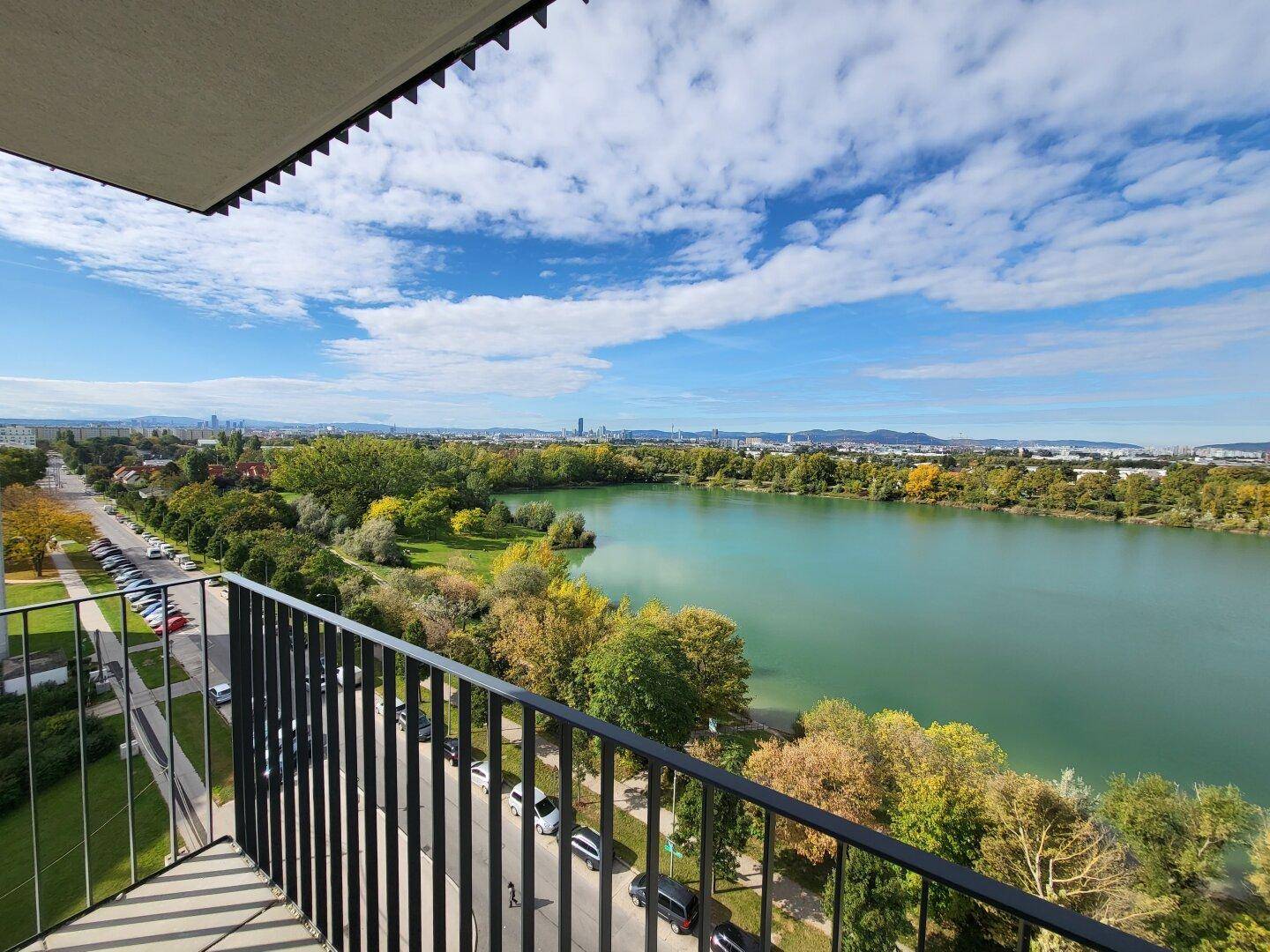 Weitläufiger Balkonblick auf den See, grüne Bäume, die Straße und die Stadt unter blauem Himmel.