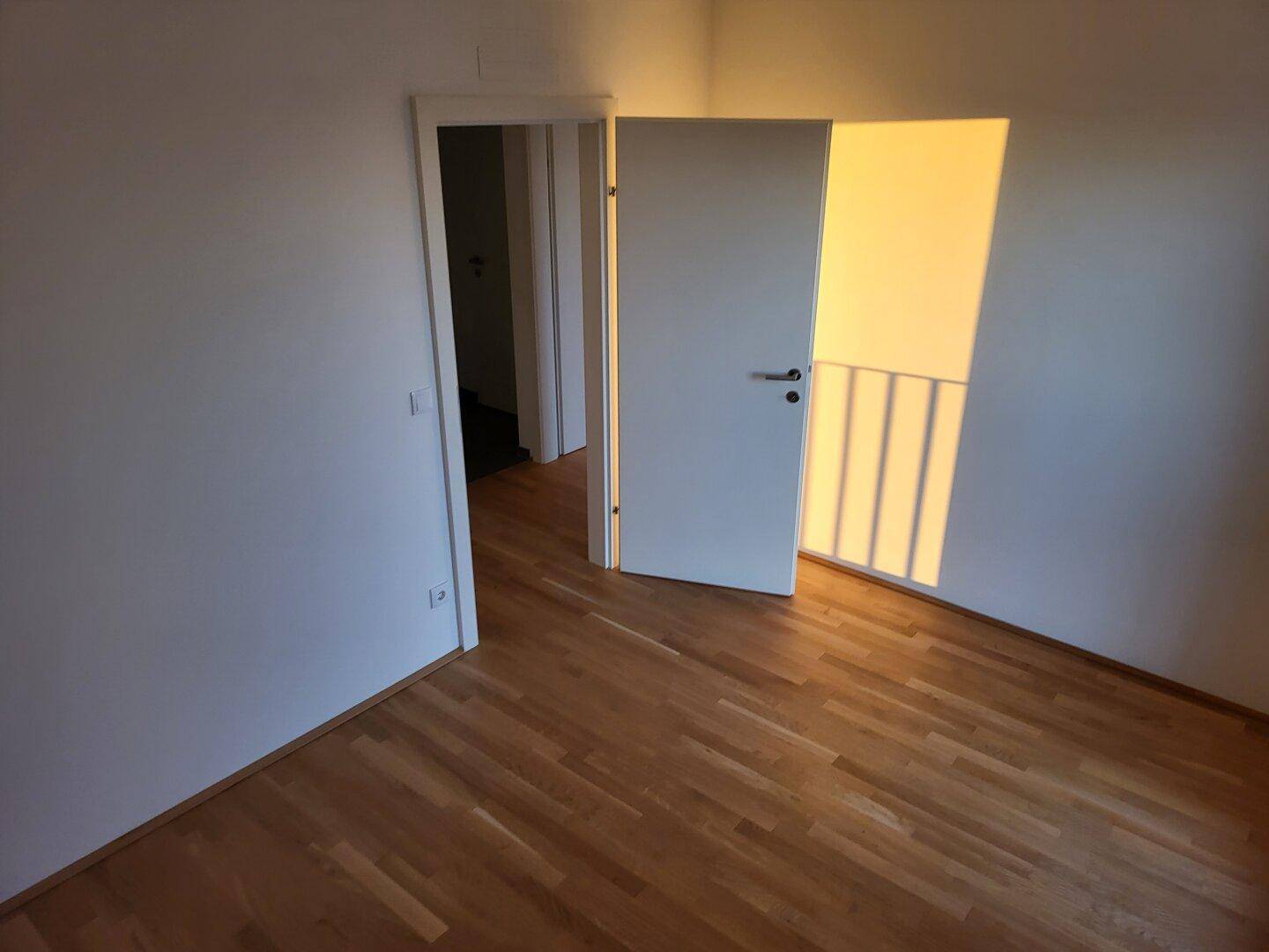 Helles Zimmer mit Parkettboden und sonnigem Lichteinfall durch die offene Tür.