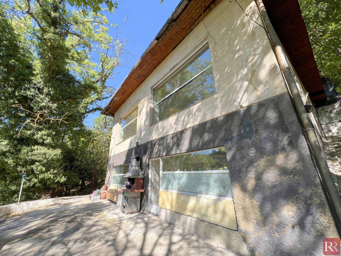 Außenbereich des Hauses mit gepflasterter Terrasse und einem gemauerten Grill, umgeben von Bäumen.