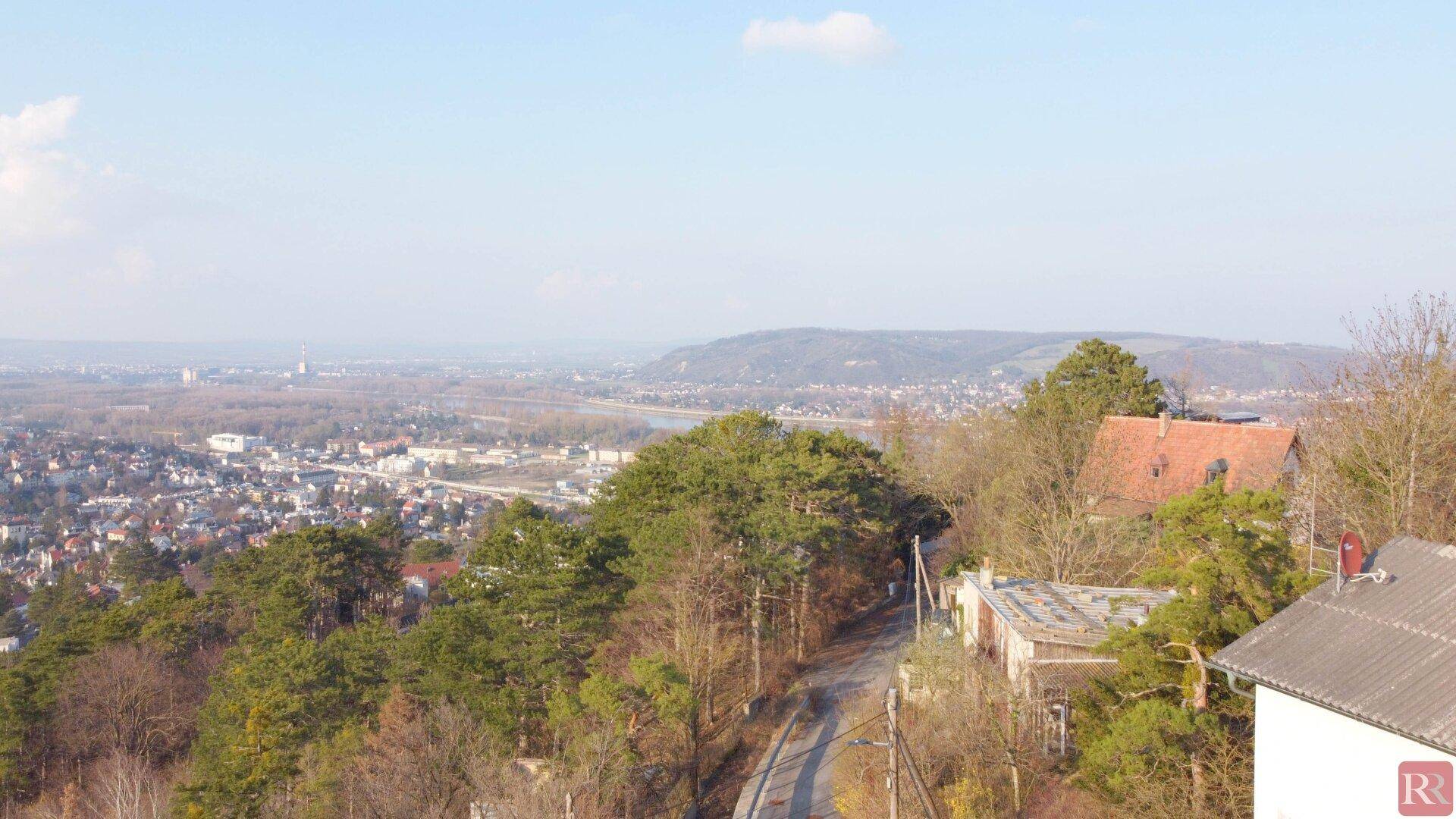 Luftaufnahme mit Blick auf eine Stadt, einen Fluss und bewaldete Hügel unter klarem Himmel.
