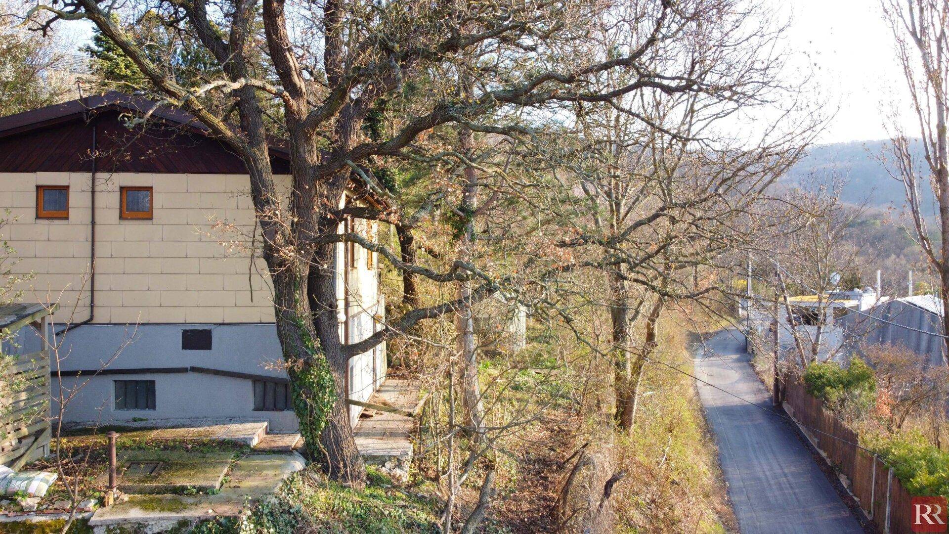 Seitenansicht eines Hauses mit einfacher Fassade, umgeben von Bäumen und einem unbefestigten Weg.