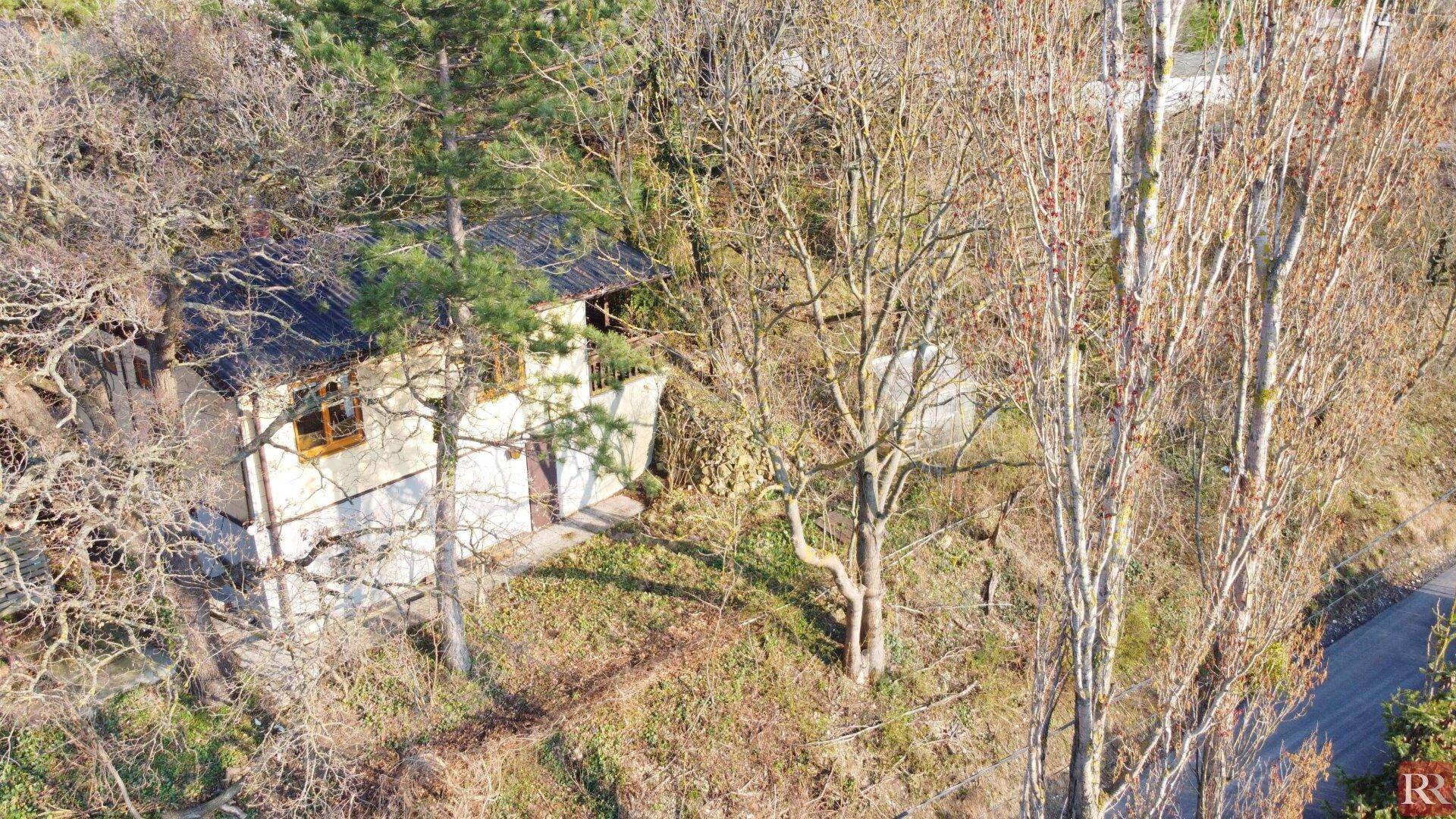 Draufsicht auf ein kleines Haus mit dunklem Dach, eingebettet in eine bewaldete Umgebung mit kahlen Bäumen.