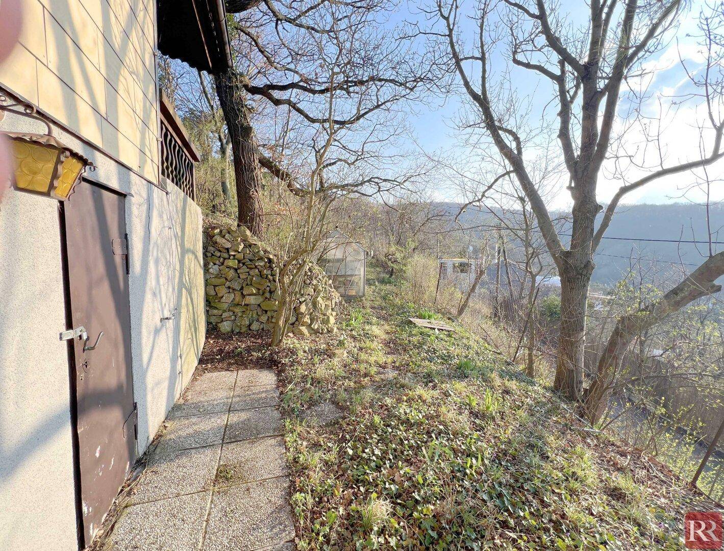 Seitenansicht eines Gebäudes mit Zugangstür, gepflastertem Weg und einer Natursteinmauer am Hang.