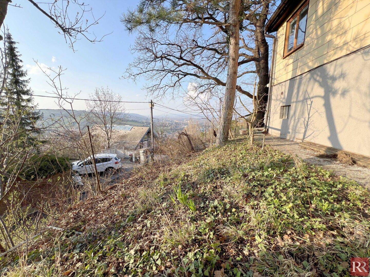 Blick vom Hanggrundstück auf die Gebäudeseite, einen geparkten Wagen auf der Straße und die weite Landschaft.