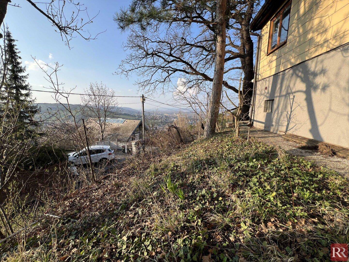 Blick vom Hanggrundstück auf die Gebäudeseite, einen geparkten Wagen auf der Straße und die weite Landschaft.