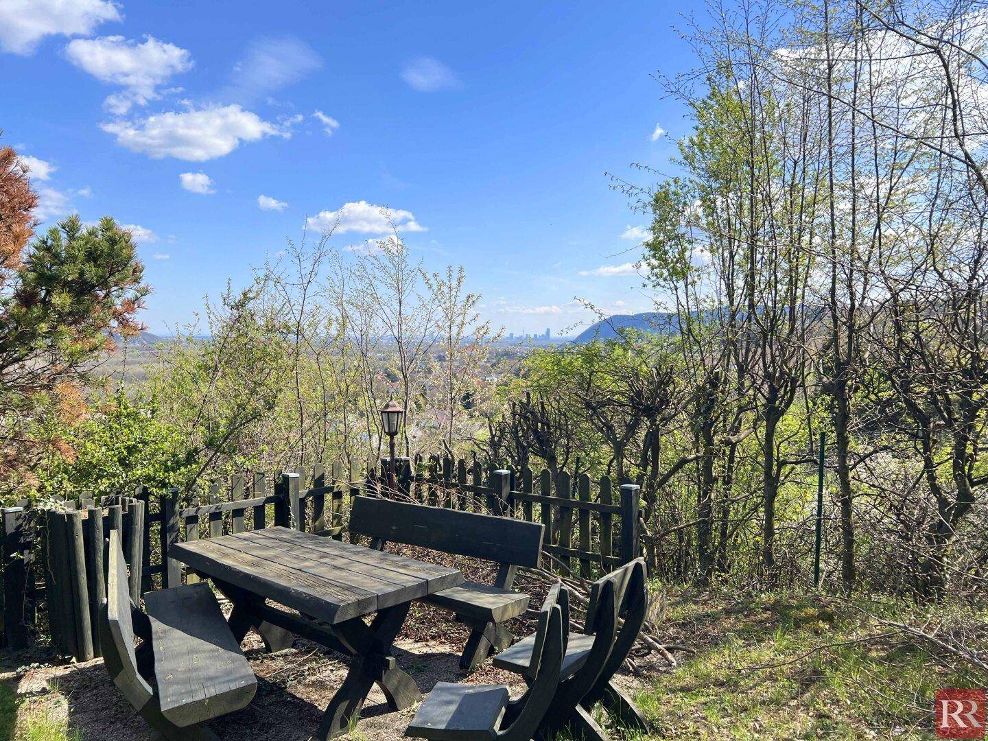 Rustikaler Sitzbereich im Freien mit Holztisch und Bänken, umgeben von Bäumen und Weitblick.