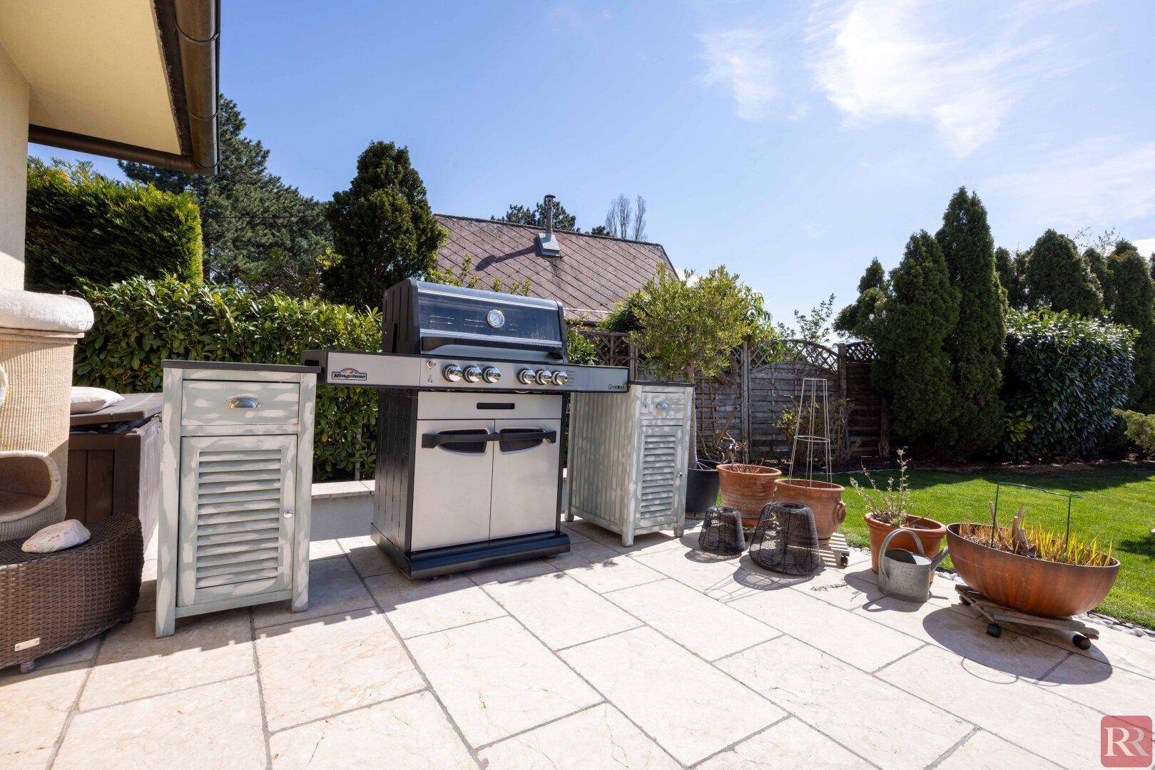 Geräumige Terrasse mit Grillbereich und verschiedenen Pflanzen in Töpfen unter blauem Himmel.