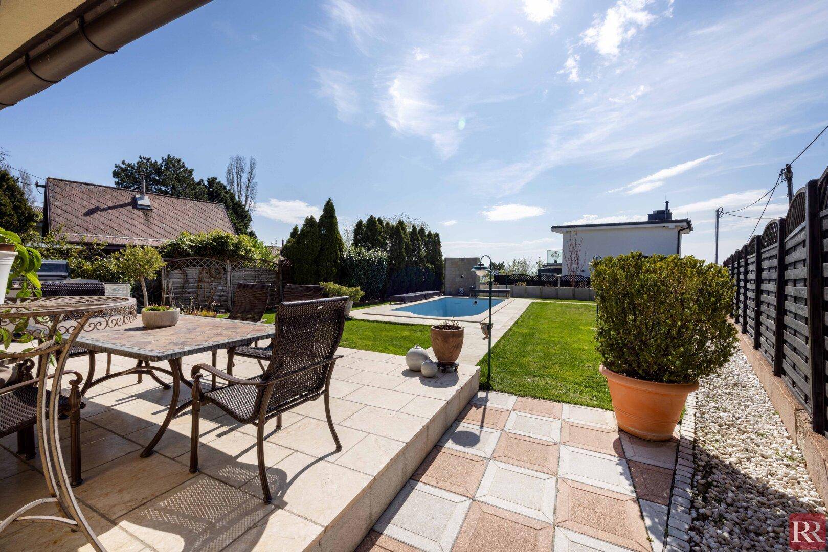 Sonniger Terrassenbereich mit Essgruppe und Blick auf den gepflegten Rasen und den Pool.