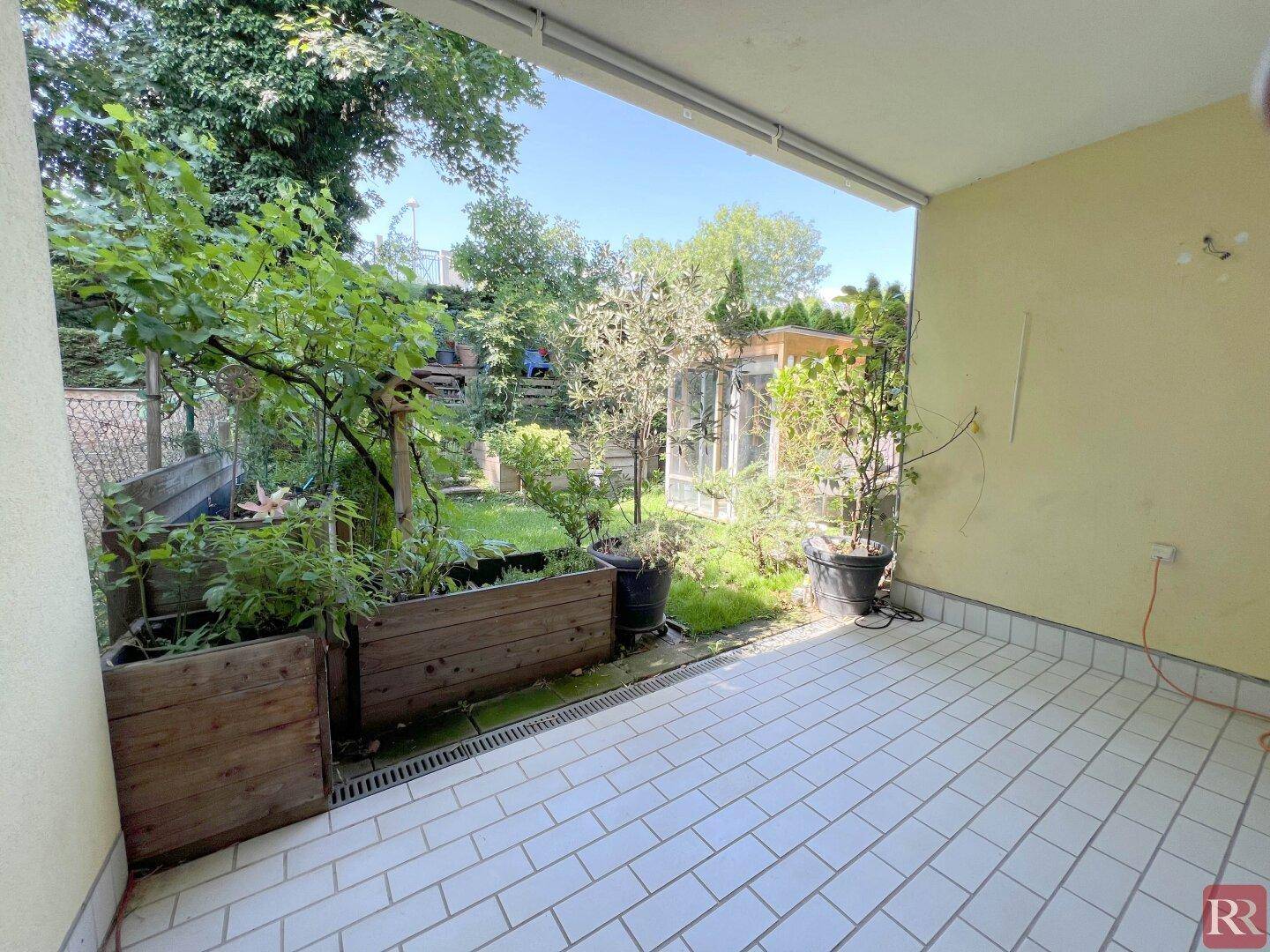 Überdachte Terrasse mit Fliesenboden, Pflanzen in Holzkästen und Blick in den grünen Garten.