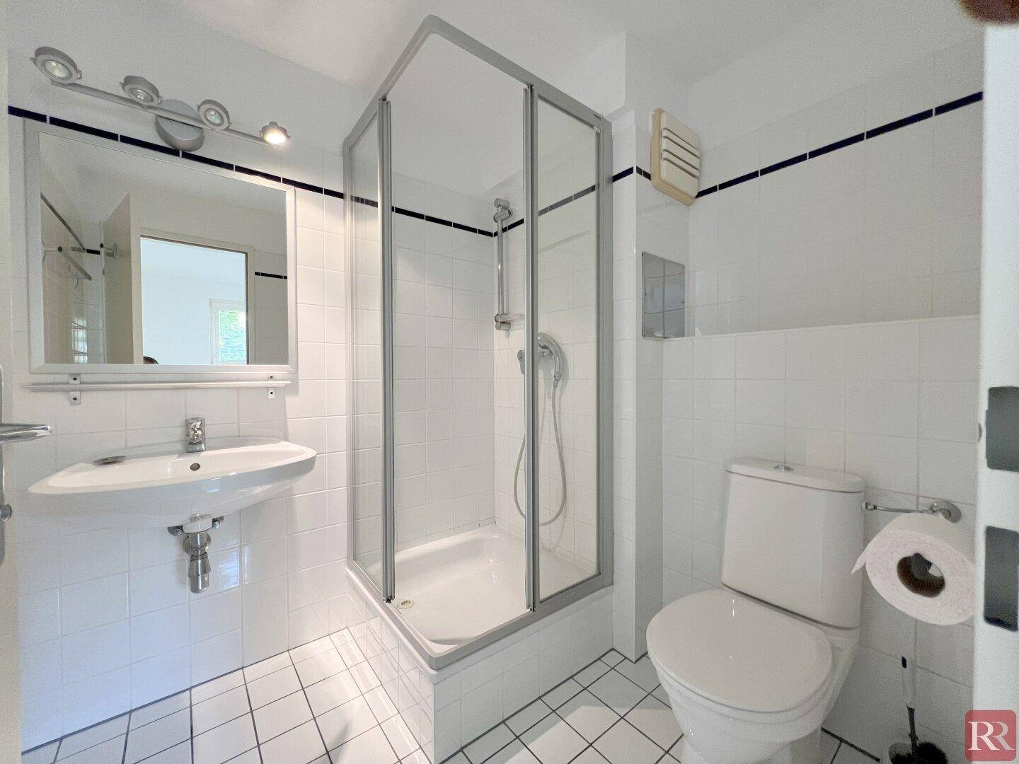 Kleines Badezimmer mit weißen Fliesen, Duschkabine, Toilette und Einzelwaschbecken.