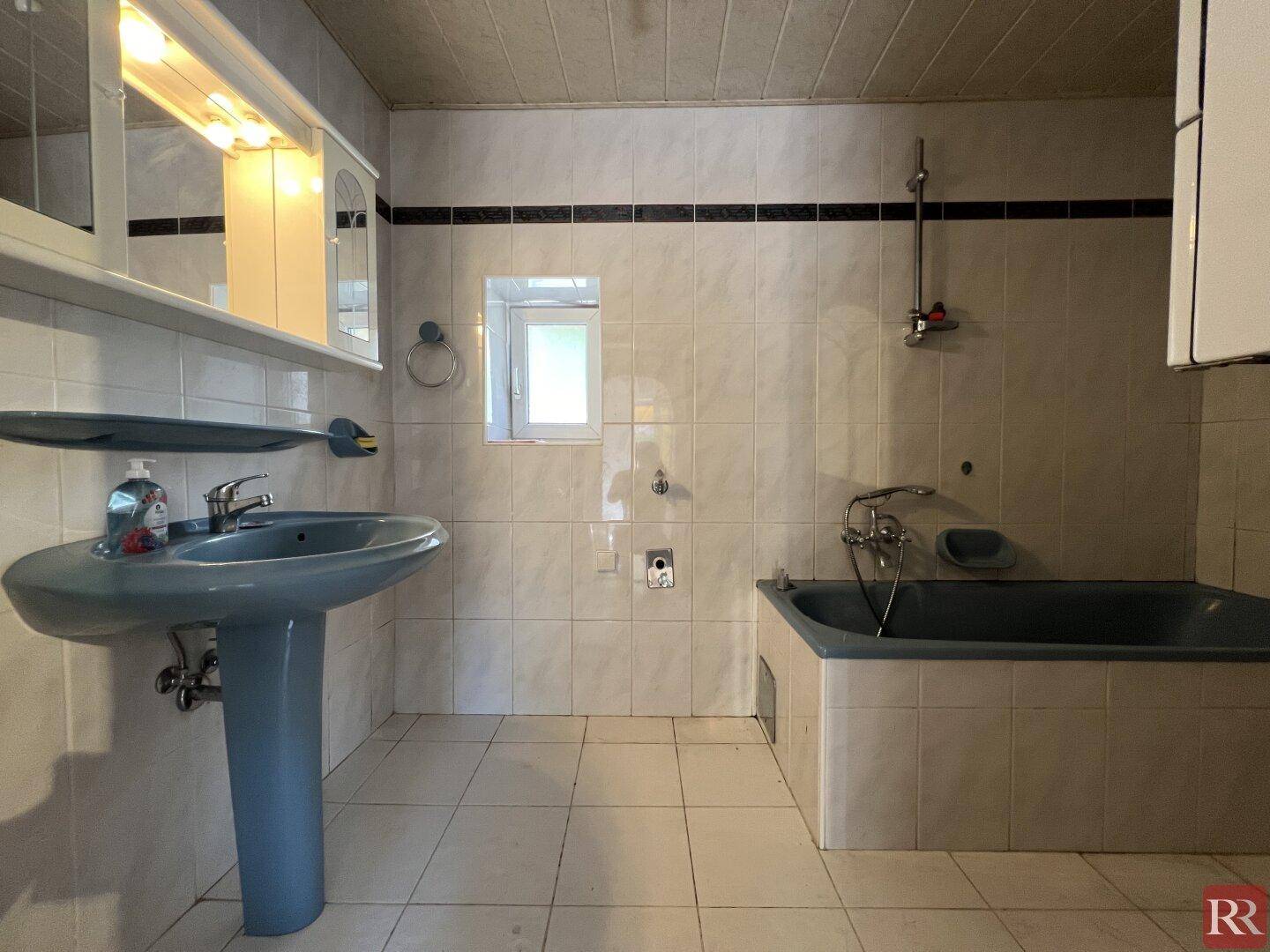Badezimmer mit blauer Badewanne, Waschbecken und weißen Wandfliesen, sowie einem kleinen Fenster.