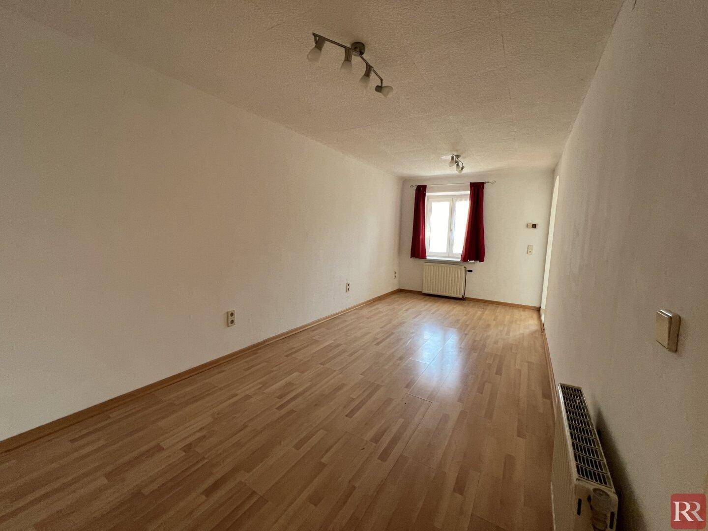 Leerer Raum mit hellem Laminatboden, weißen Wänden und einem Fenster mit roten Vorhängen.