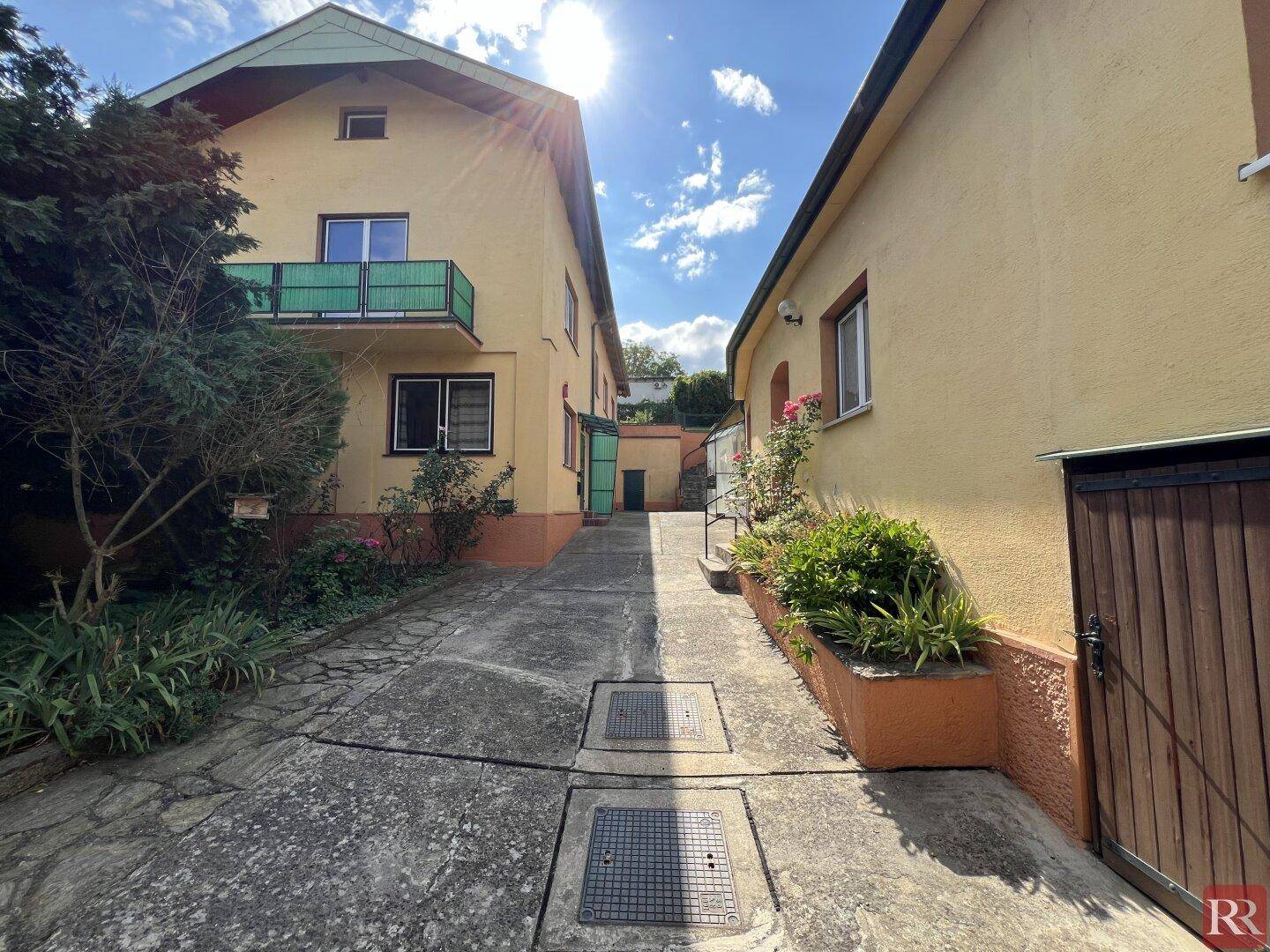 Gelbes Wohnhaus mit grünem Balkon und angrenzendem Gebäude, verbunden durch eine gepflasterte Zufahrt.