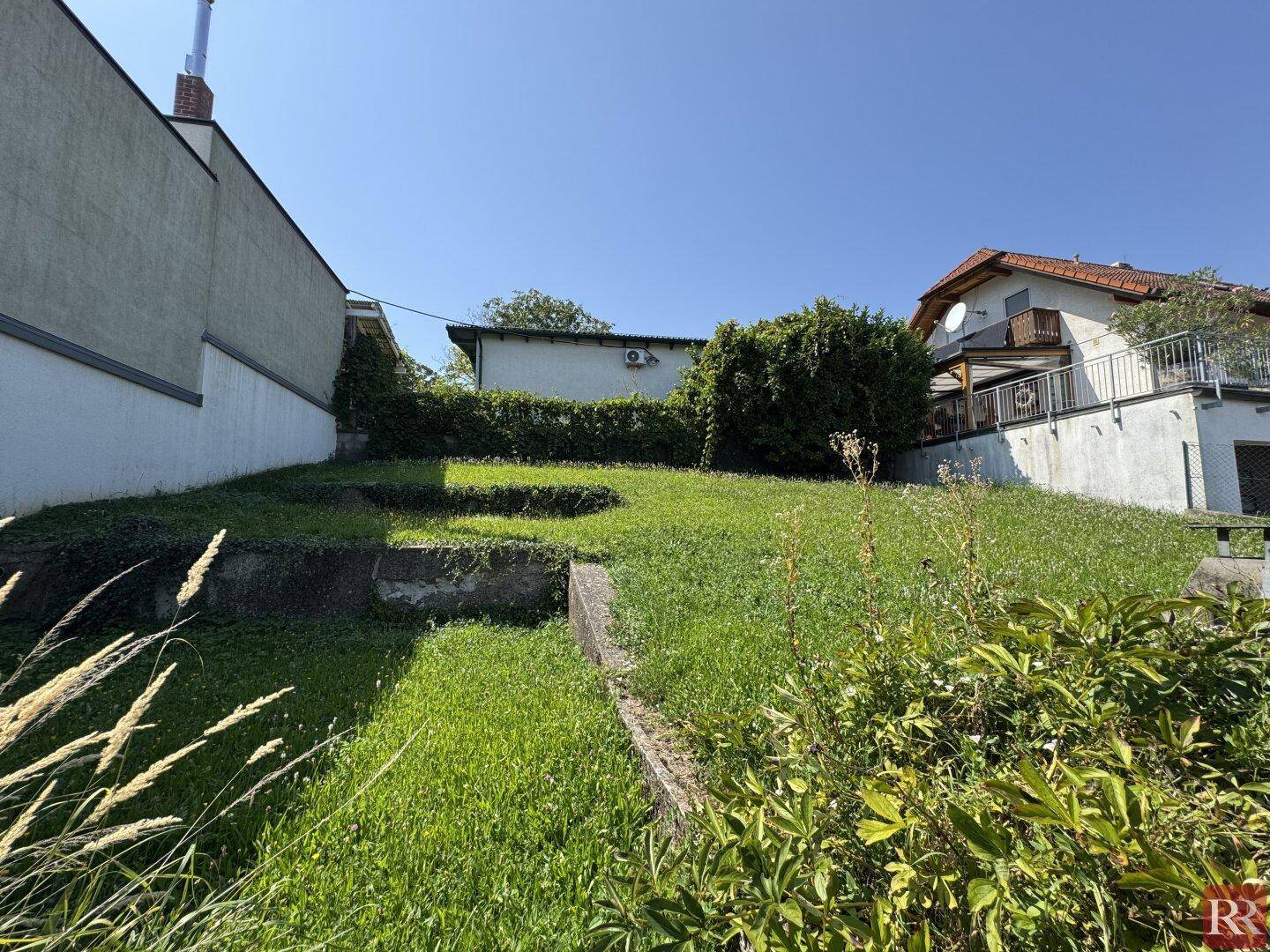 Terrassierter Gartenbereich mit grüner Wiese und Betonmauern unter strahlend blauem Himmel.