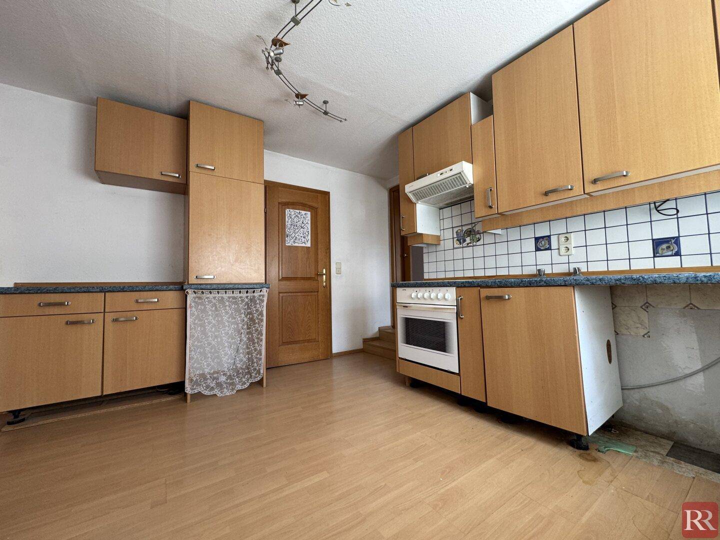 Küche mit hellen Holzschränken, Herd und Spüle, sowie einem Fenster und unterschiedlichen Bodenbelägen.
