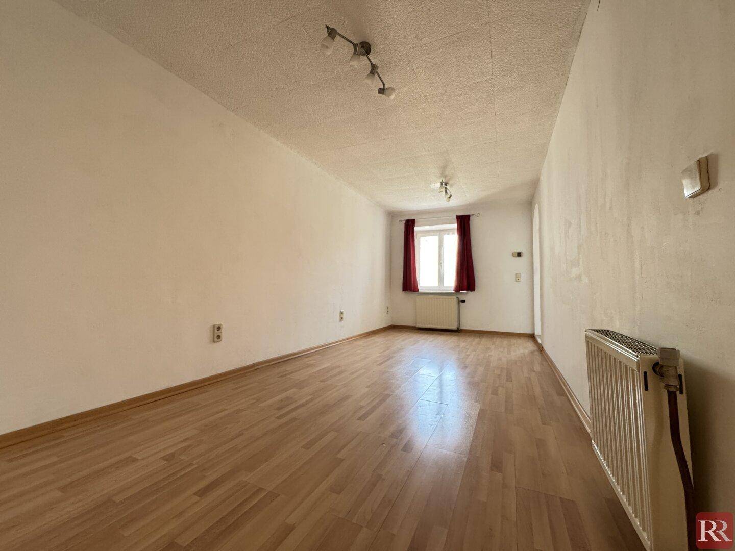 Leerer Raum mit hellem Laminatboden, weißen Wänden und einem Fenster mit roten Vorhängen.