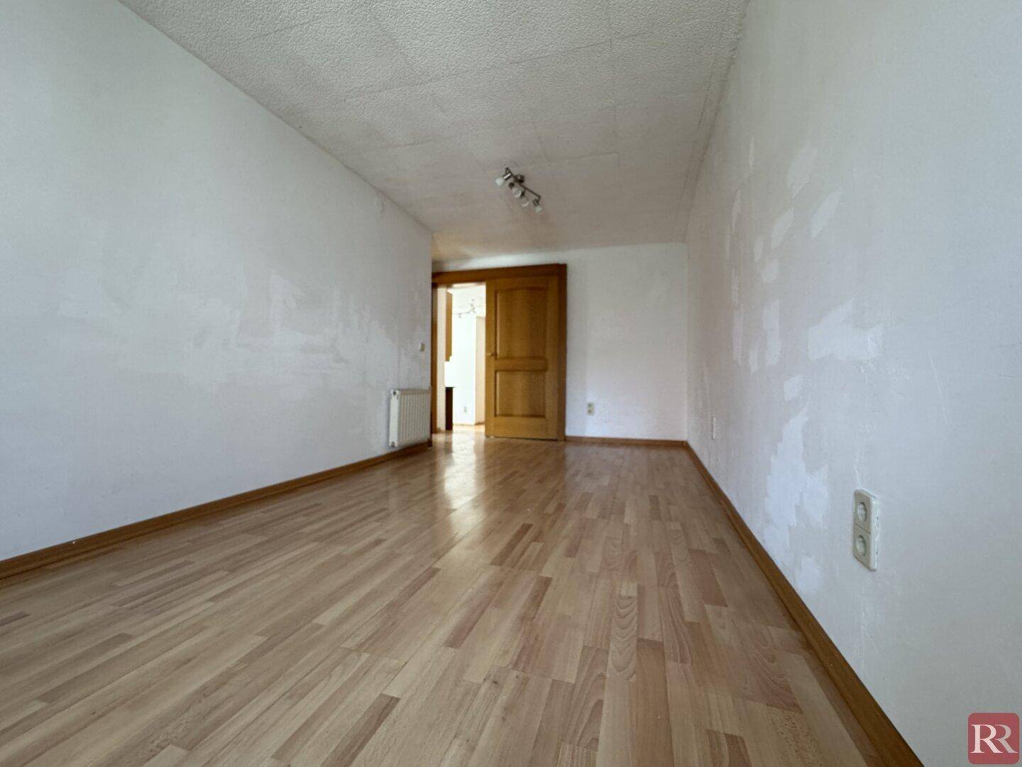 Heller, leerer Raum mit Laminatboden und weißen Wänden, der zu einem weiteren Zimmer führt.