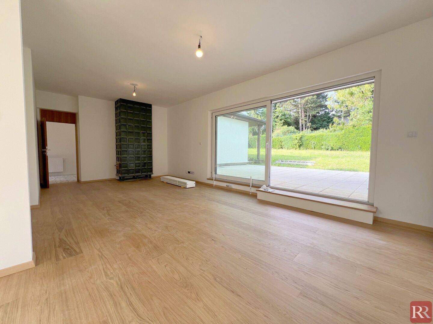 Geräumiges Wohnzimmer mit Holzboden, Kachelofen und Zugang zur Terrasse.