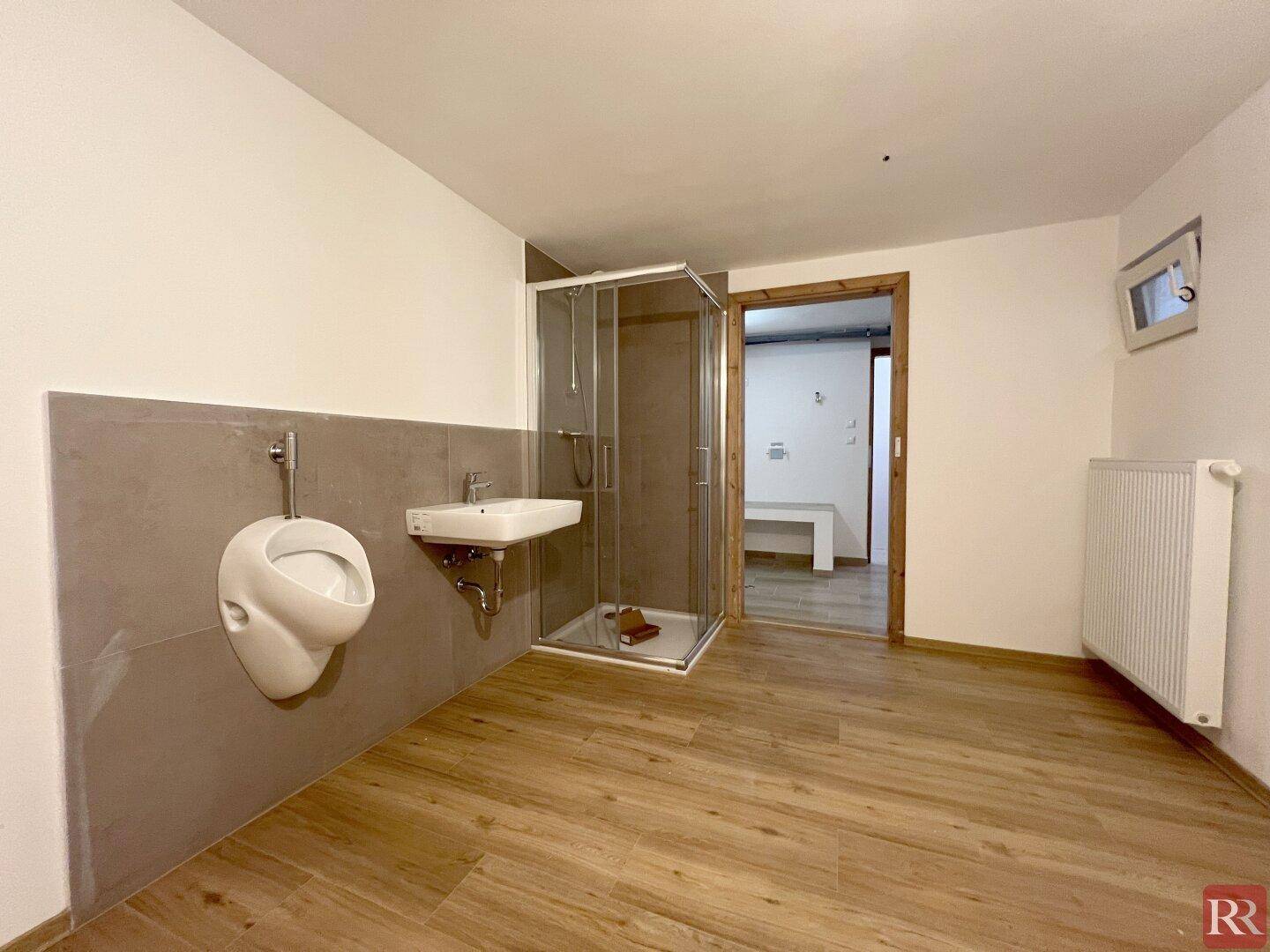 Badezimmer mit Urinal, Waschbecken und Duschkabine, sowie Holzoptik-Boden.