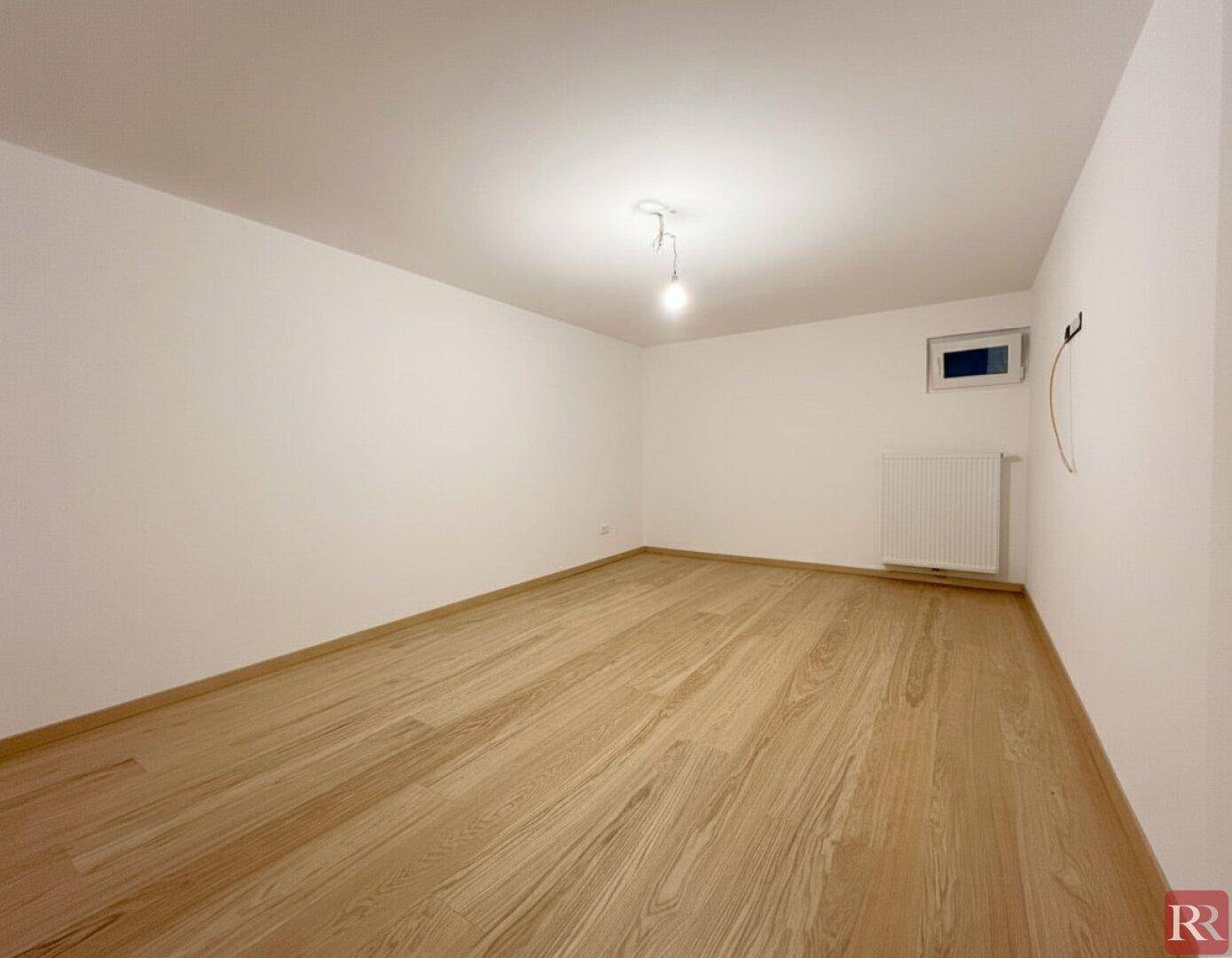 Leeres Zimmer mit hellem Holzboden und kleinem Fenster.
