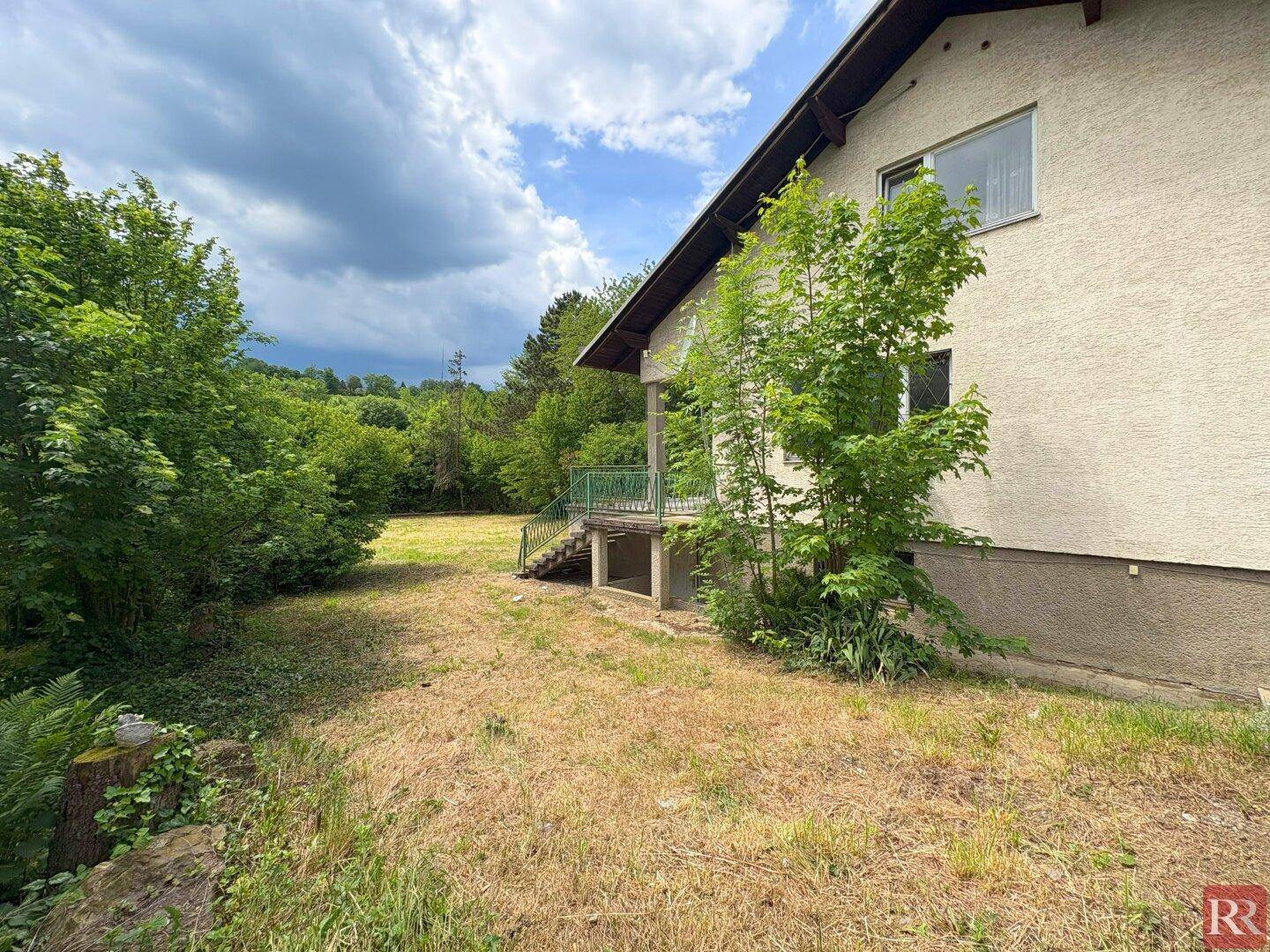 Seitenansicht des Hauses mit Balkon und Treppe, umgeben von üppigem Grün und Garten.