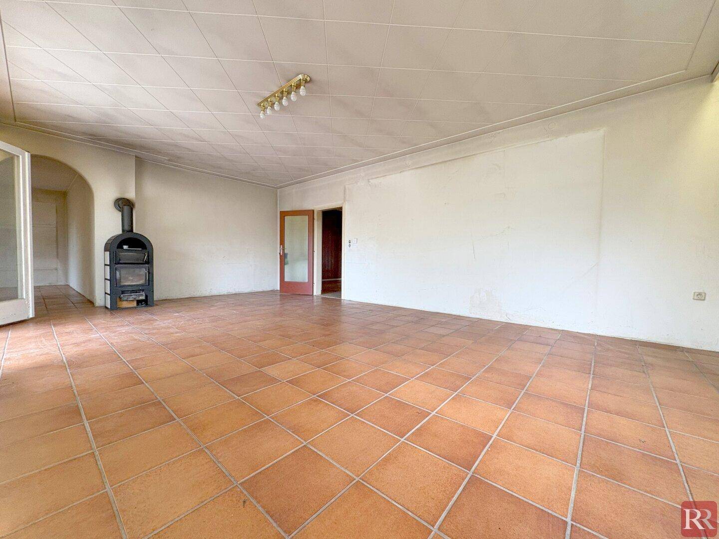 Geräumiges Wohnzimmer mit Kaminofen, Terrakottafliesen und Blick in den Flur.