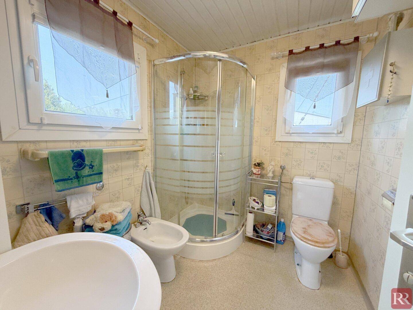 Funktionales Badezimmer mit Duschkabine, WC, Bidet und zwei Fenstern für Tageslicht.