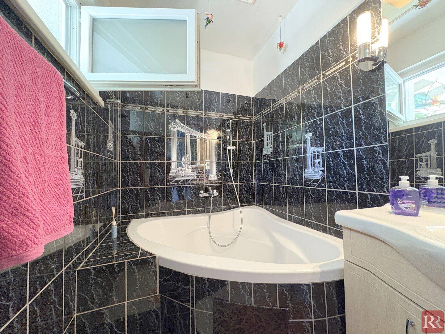 Badezimmer mit dunklen Fliesen, Eckbadewanne und kleinem Fenster für natürliche Belüftung.