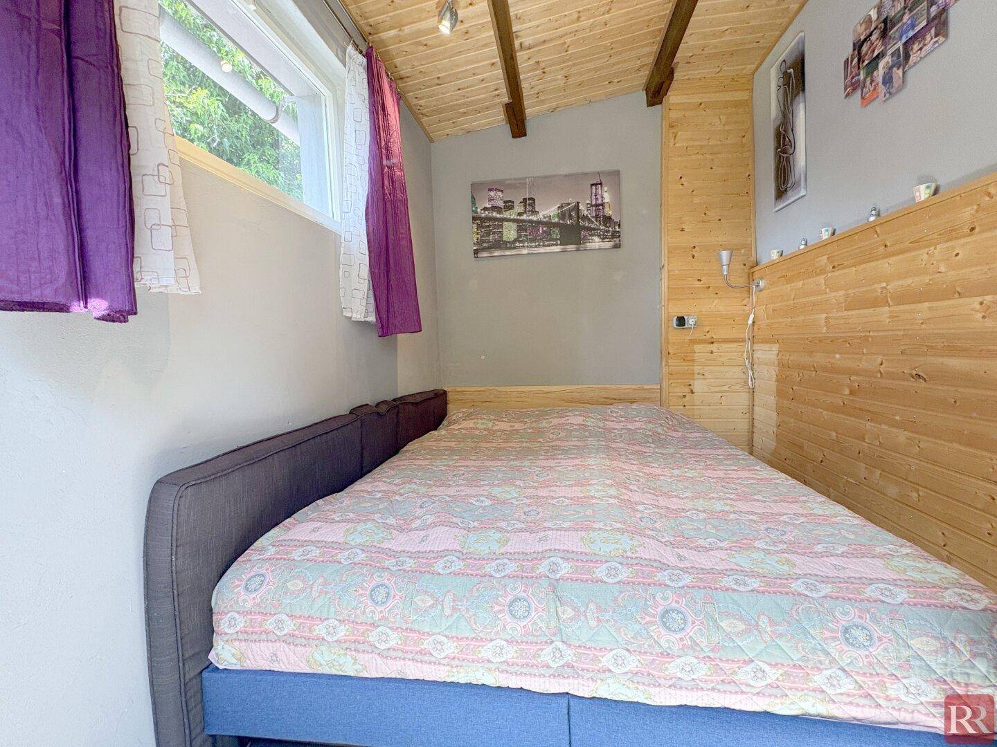 Schlafzimmer mit Holzwänden, Holzbalkendecke und einem Bett unter dem Fenster, das eine gemütliche Atmosphäre schafft.
