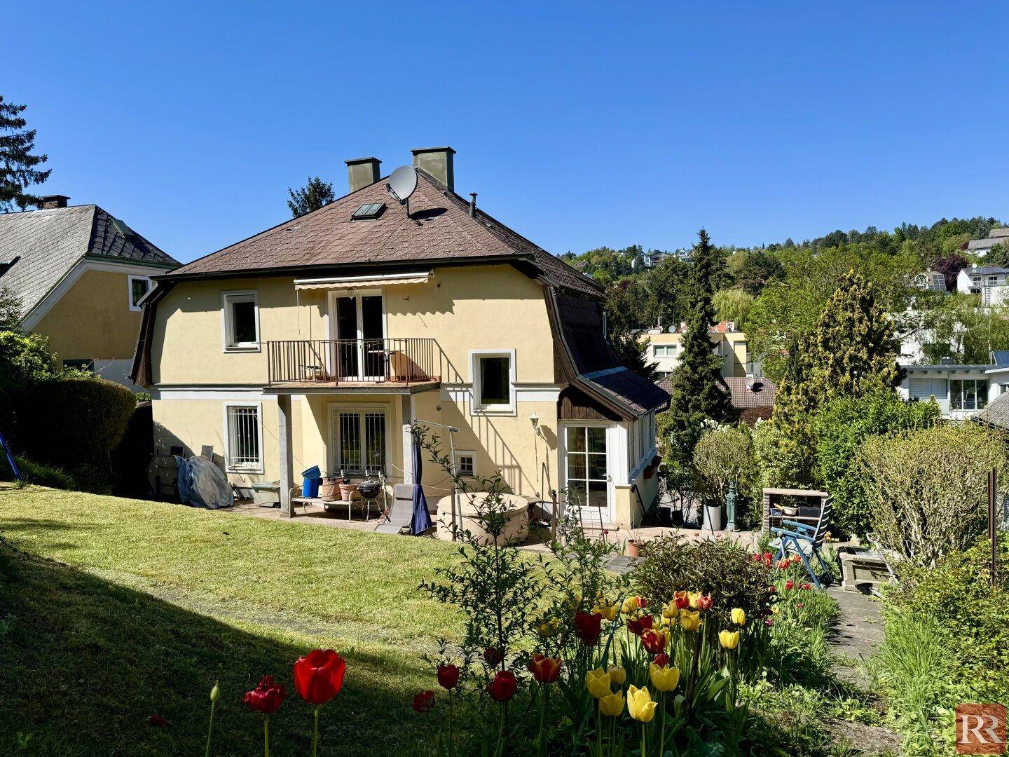 Gartenbereich hinter dem Haus mit blühenden Blumen, Rasenfläche und Blick auf die umliegende grüne Landschaft.