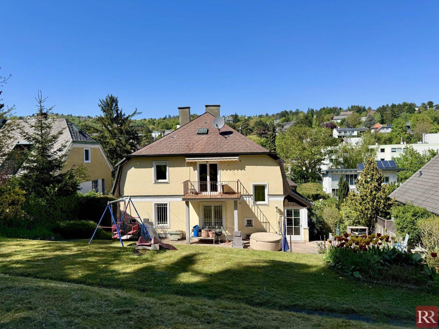 Hinterer Gartenbereich des Hauses mit Rasenfläche, Schaukel und Rutsche für Kinder unter strahlendem Himmel.