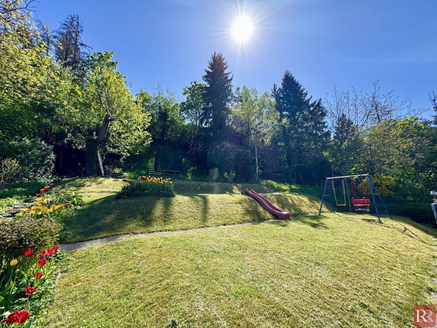 Großer, sonniger Garten mit Rasenflächen, Rutsche und Schaukel, umgeben von Bäumen und viel Grün.