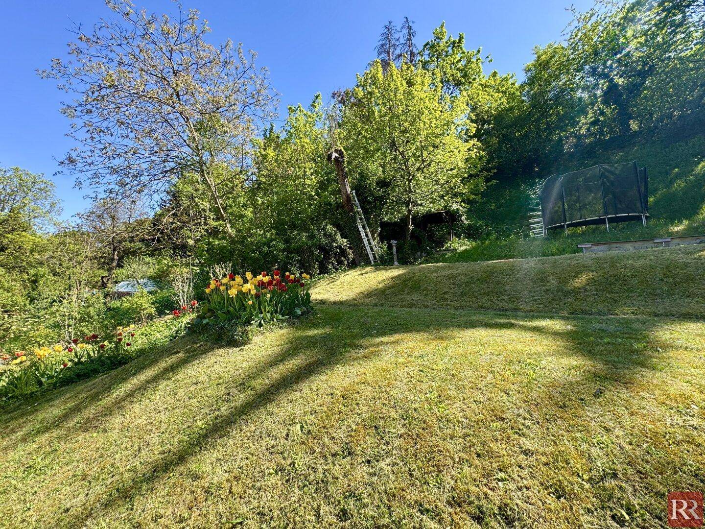 Weitläufiger Gartenbereich mit Rasenfläche, Blumenbeeten und einem Trampolin im Hintergrund unter blauem Himmel.