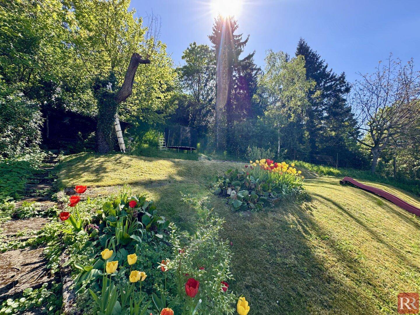 Sonniger Garten mit bunten Blumenbeeten, Rasenfläche und einer Rutsche, umgeben von hohen Bäumen.