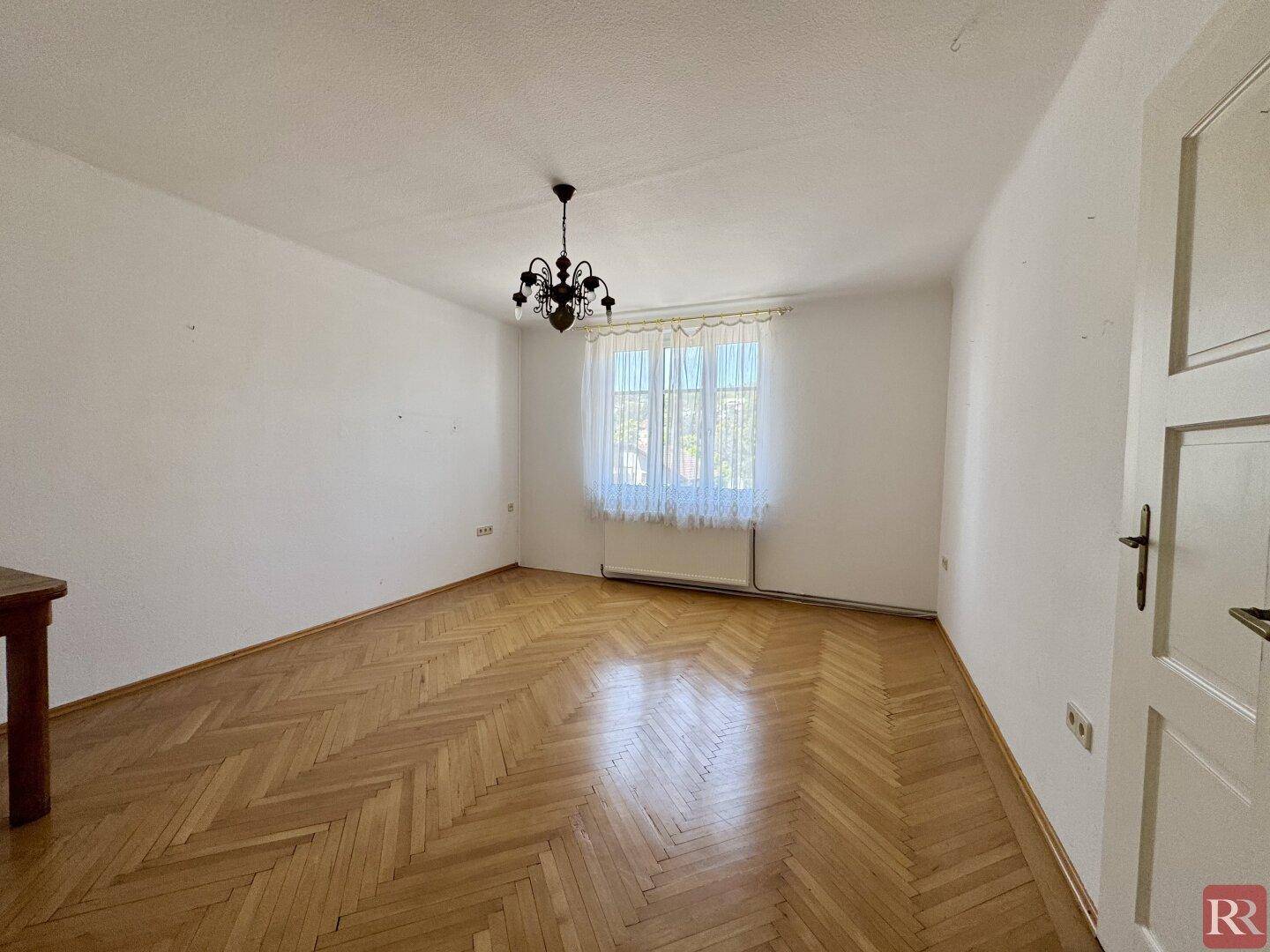 Lichtdurchflutetes Wohnzimmer mit Fischgrätparkett, einem Fenster mit Vorhängen und einem Heizkörper.