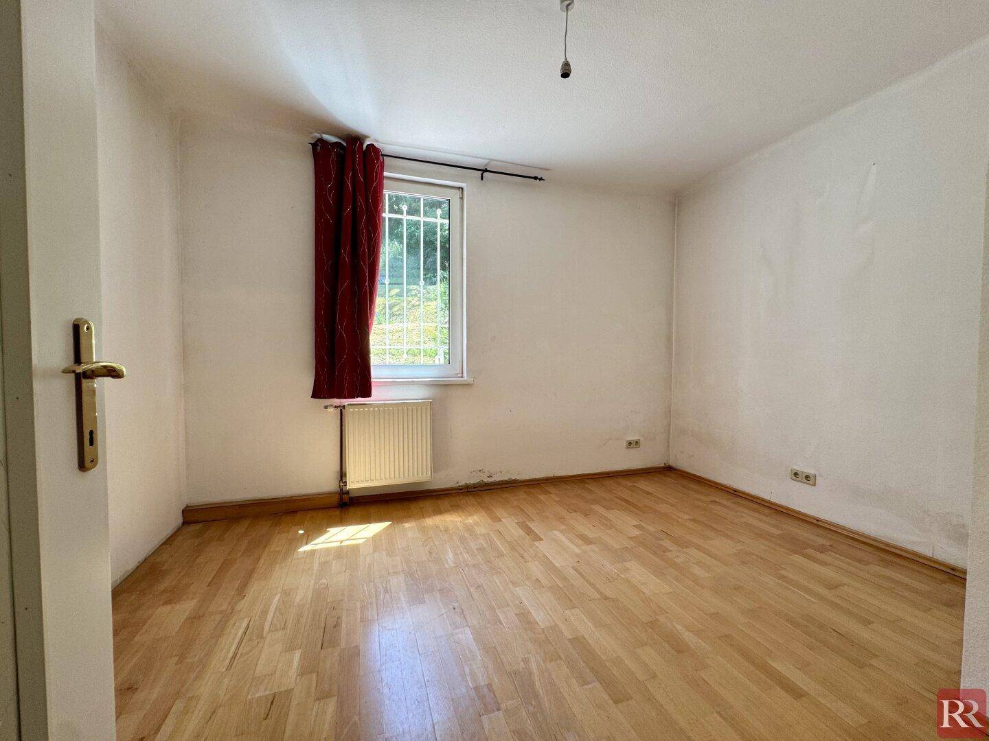 Kleines Schlafzimmer mit Holzboden, einem Fenster mit Gitter und einem roten Vorhang.