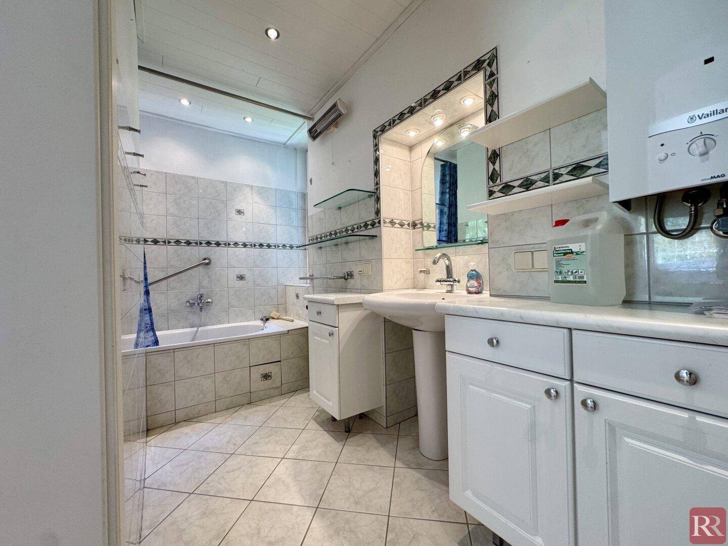 Gefliestes Badezimmer mit Badewanne, Waschtisch und einem großen Spiegel mit Beleuchtung.