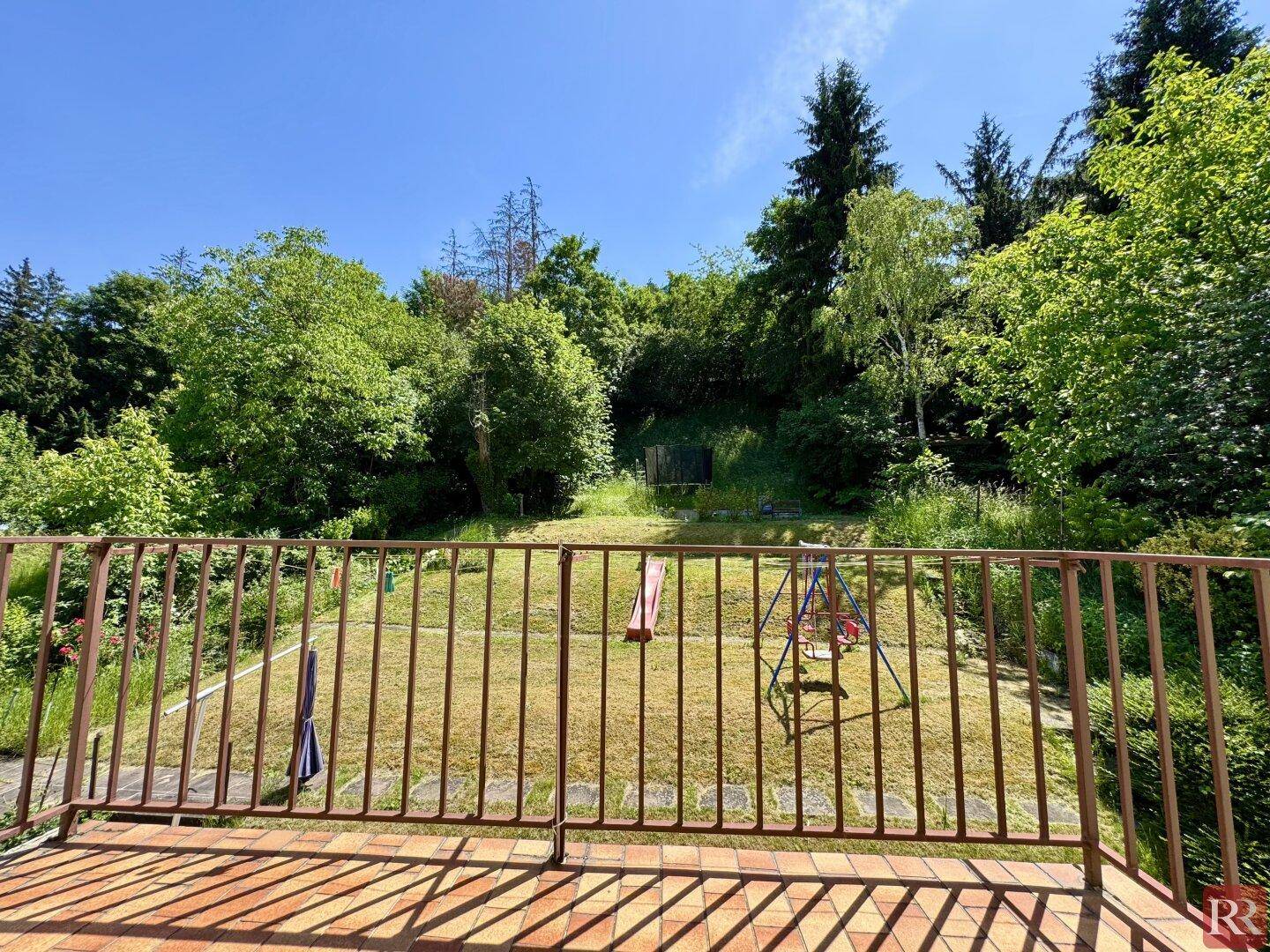 Blick vom Balkon auf den weitläufigen Garten mit Rasenfläche und Spielgeräten unter blauem Himmel.