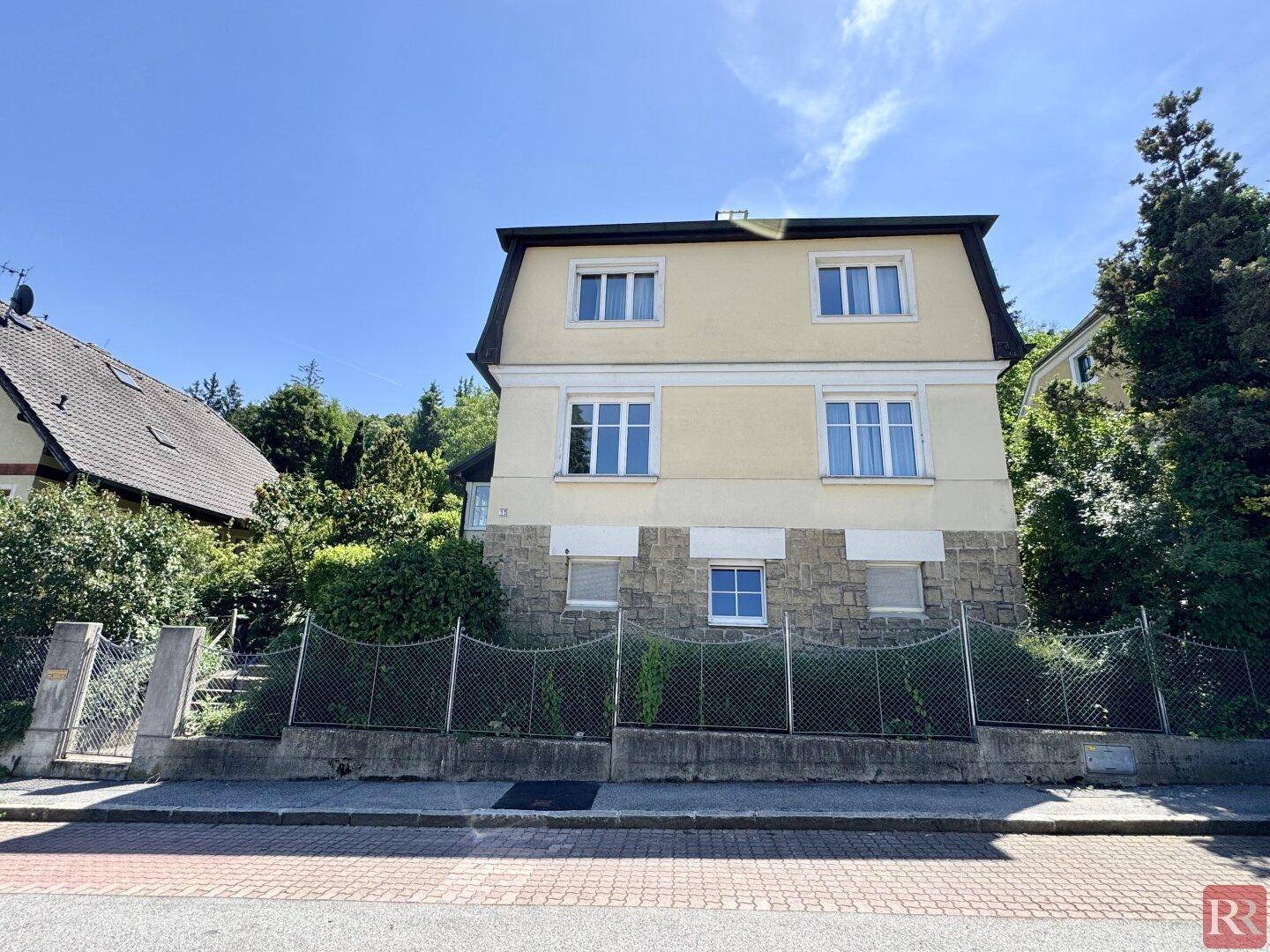 Vorderansicht des Hauses von der Straße aus mit gelber Fassade, mehreren Fenstern und einem Zaun.