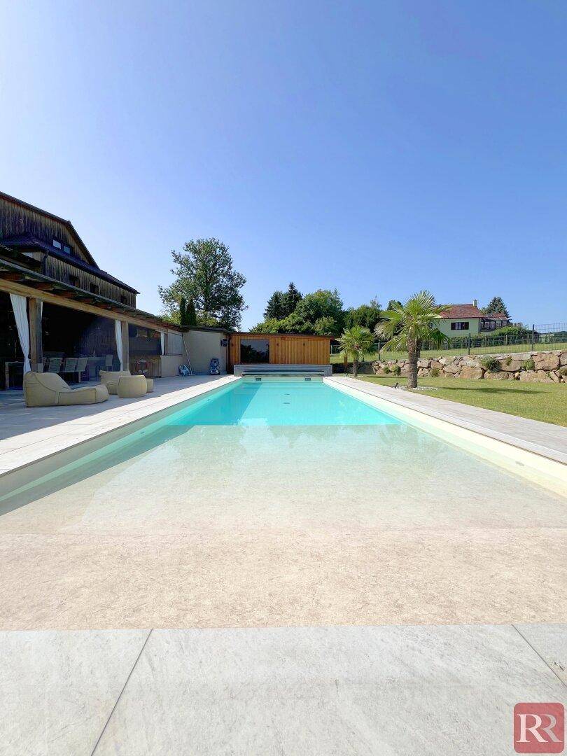 Weitläufiger Poolbereich mit flachem Einstieg, umgeben von Rasenflächen und einem Holzgebäude.