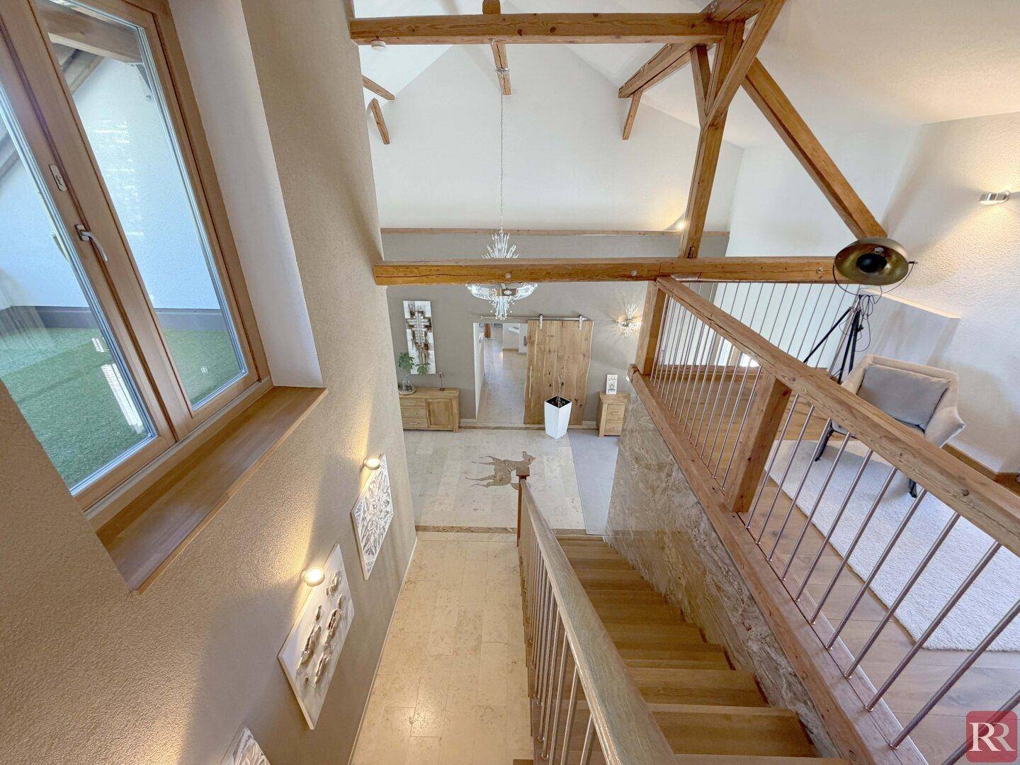 Treppenhaus mit Holzgeländer und Blick auf den hohen Eingangsbereich mit Holzbalken und Kronleuchter.