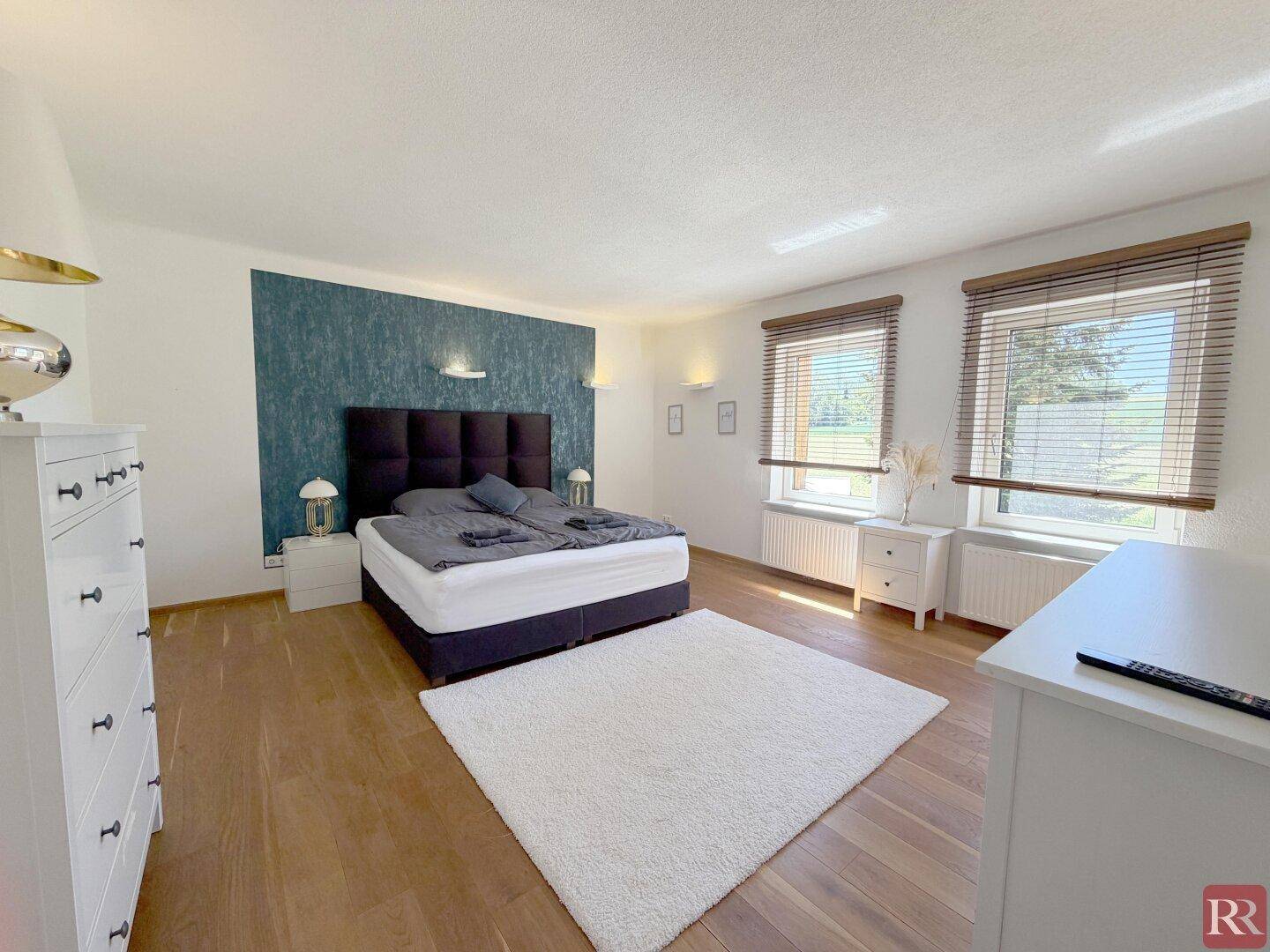 Helles Schlafzimmer mit Holzboden, großem Bett und zwei Fenstern, die viel Tageslicht hereinlassen.