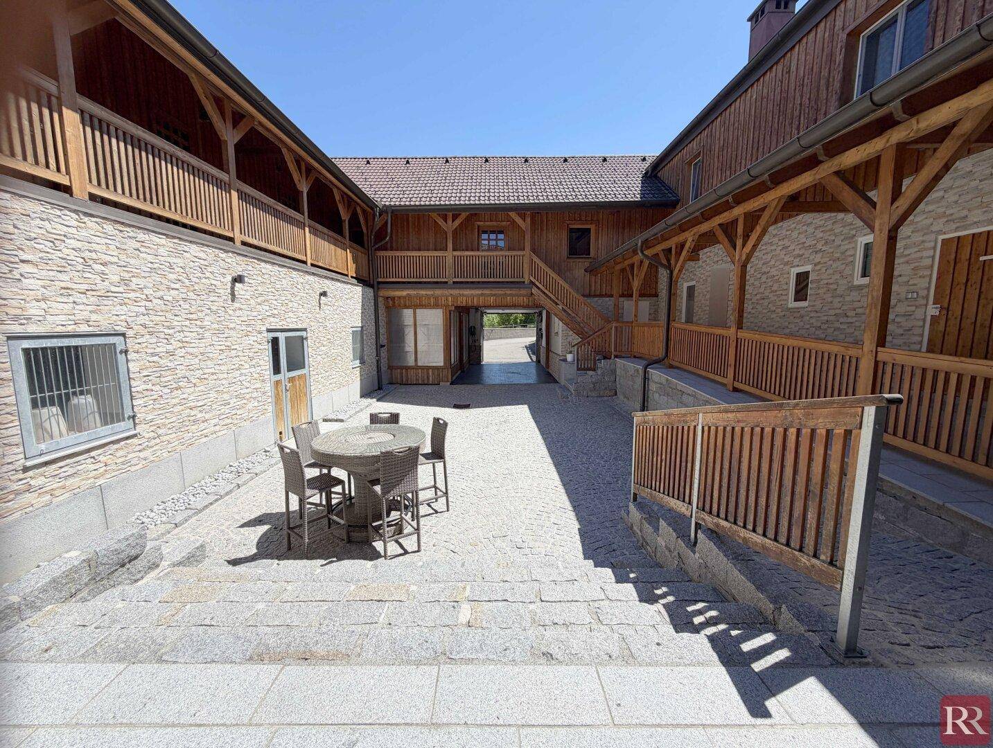 Innenhof mit Steinfliesen, Sitzgelegenheiten und rustikalen Holzfassaden der umliegenden Gebäude.