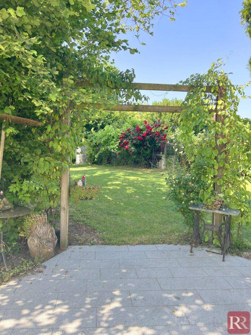 Idyllischer Gartenbereich mit bewachsenem Torbogen, Rasenfläche und blühenden Rosenbüschen.
